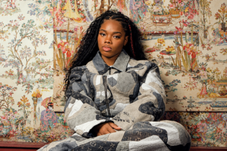 Cristale sits in a grey and cream patchwork monogram denim set, with long braids, against ornate hand-painted floral wallpaper.