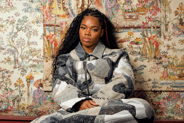 Cristale sits in a grey and cream patchwork monogram denim set, with long braids, against ornate hand-painted floral wallpaper.