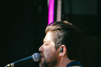 Dave Polo sings into a microphone while playing a wooden ukulele on stage, wearing a blue linen shirt, shell bracelets and an orange Surfestival wristband, with a guitarist behind him.