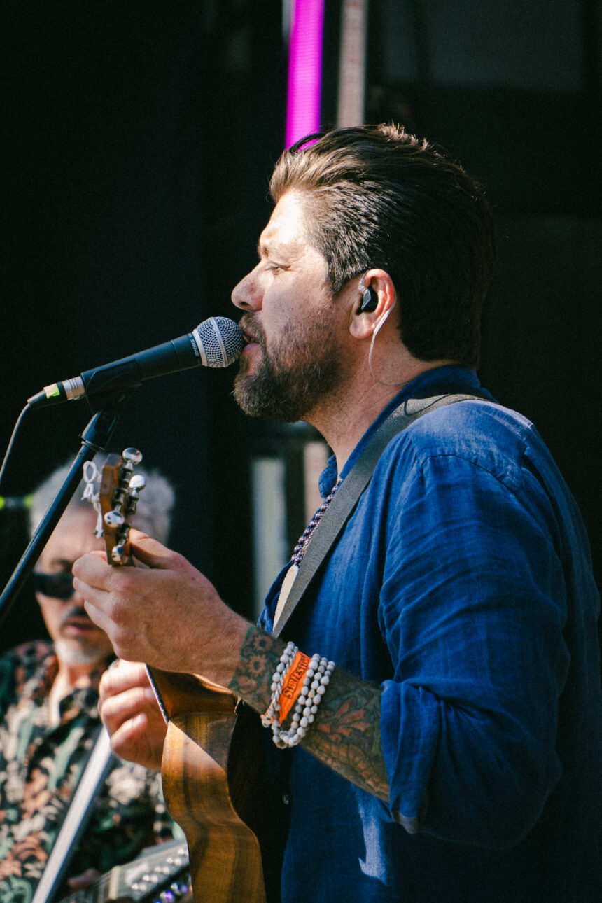 Dave Polo sings into a microphone while playing a wooden ukulele on stage, wearing a blue linen shirt, shell bracelets and an orange Surfestival wristband, with a guitarist behind him.