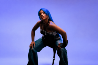 Doechii crouches against a purple-lit backdrop in a long electric-blue wig, a black leather corset top with an ornate silver trim, baggy jeans, buckled fingerless gloves and long silver nails.