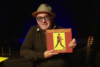 Elvis Costello, in a straw fedora and tinted glasses, holds up the red and yellow 'My Aim Is True' box set on a dimly lit stage, with an acoustic guitar beside him.