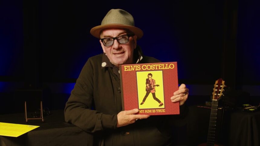 Elvis Costello, in a straw fedora and tinted glasses, holds up the red and yellow 'My Aim Is True' box set on a dimly lit stage, with an acoustic guitar beside him.