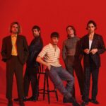 The five members of Fontaines D.C. pose against a bold red backdrop, one seated on a stool at the centre, the others standing in tailored suits, a striped polo and a knit jumper.