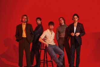 The five members of Fontaines D.C. pose against a bold red backdrop, one seated on a stool at the centre, the others standing in tailored suits, a striped polo and a knit jumper.