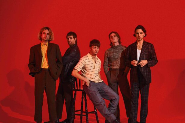 The five members of Fontaines D.C. pose against a bold red backdrop, one seated on a stool at the centre, the others standing in tailored suits, a striped polo and a knit jumper.