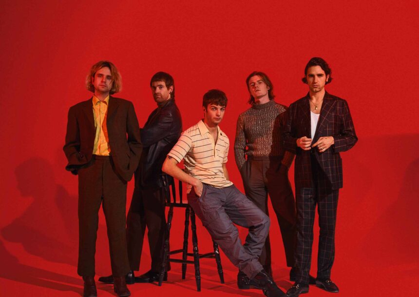The five members of Fontaines D.C. pose against a bold red backdrop, one seated on a stool at the centre, the others standing in tailored suits, a striped polo and a knit jumper.
