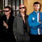 Fontaines D.C. pose in a row against a weathered yellow wall with barred windows, dressed in dark jackets, sunglasses and a blue tracksuit top, ahead of new single 'Tongue'.