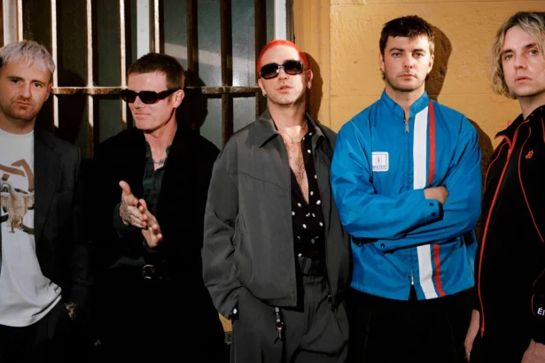 Fontaines D.C. pose in a row against a weathered yellow wall with barred windows, dressed in dark jackets, sunglasses and a blue tracksuit top, ahead of new single 'Tongue'.