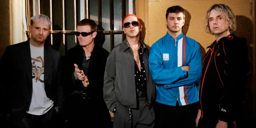 Fontaines D.C. pose in a row against a weathered yellow wall with barred windows, dressed in dark jackets, sunglasses and a blue tracksuit top, ahead of new single 'Tongue'.