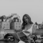 Singer-songwriter Jesabel reads a copy of Le Monde by the Seine, with a Paris bridge and historic rooftops behind her, in a black-and-white promotional photo for her single ‘Men of Paris.’