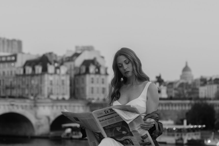 Singer-songwriter Jesabel reads a copy of Le Monde by the Seine, with a Paris bridge and historic rooftops behind her, in a black-and-white promotional photo for her single ‘Men of Paris.’