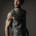 Manu Francois looks to the side in a sleeveless floral-print vest, tattoos visible on his arm, against a dark brick wall, in a promotional portrait for his single ‘Oncoming Life.’