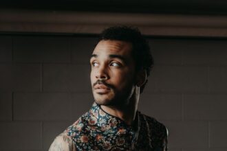 Manu Francois looks to the side in a sleeveless floral-print vest, tattoos visible on his arm, against a dark brick wall, in a promotional portrait for his single ‘Oncoming Life.’