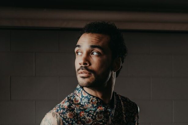 Manu Francois looks to the side in a sleeveless floral-print vest, tattoos visible on his arm, against a dark brick wall, in a promotional portrait for his single ‘Oncoming Life.’