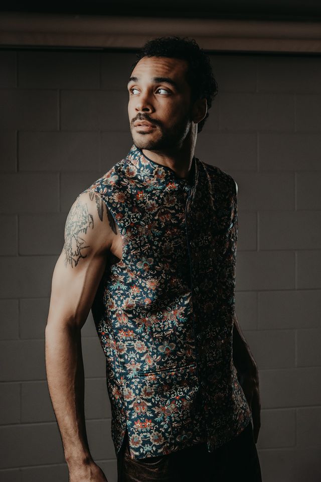 Manu Francois looks to the side in a sleeveless floral-print vest, tattoos visible on his arm, against a dark brick wall, in a promotional portrait for his single ‘Oncoming Life.’