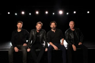 Nickelback’s four members sit side by side on the edge of a dark stage, dressed in black, under a row of white stage lights.