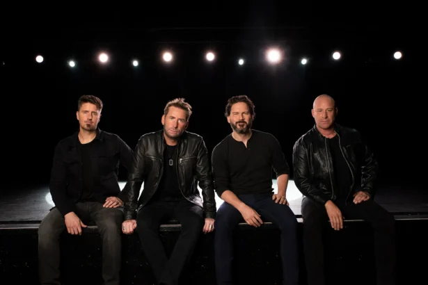 Nickelback’s four members sit side by side on the edge of a dark stage, dressed in black, under a row of white stage lights.
