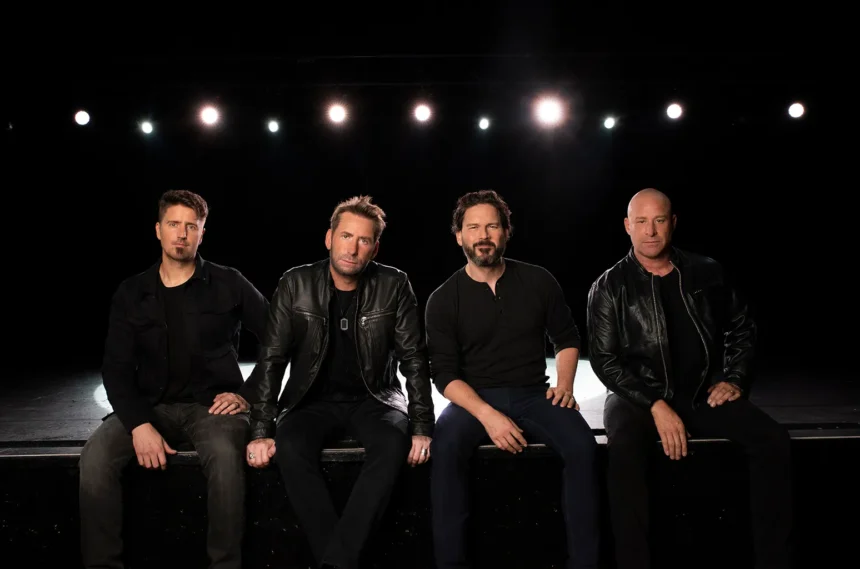 Nickelback’s four members sit side by side on the edge of a dark stage, dressed in black, under a row of white stage lights.