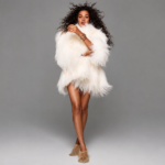 Olivia Dean stands against a plain grey backdrop in a fluffy white shaggy fur coat and gold fringed boots, with voluminous curly hair and stacked gold bangles.