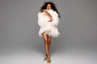 Olivia Dean stands against a plain grey backdrop in a fluffy white shaggy fur coat and gold fringed boots, with voluminous curly hair and stacked gold bangles.
