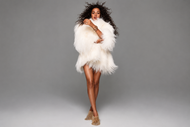 Olivia Dean stands against a plain grey backdrop in a fluffy white shaggy fur coat and gold fringed boots, with voluminous curly hair and stacked gold bangles.