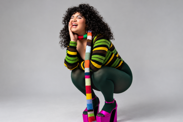 Remi Wolf crouches and laughs against a plain grey backdrop, wearing a green-striped knit top, dark green tights, hot pink platform heels and a long multicoloured striped scarf.