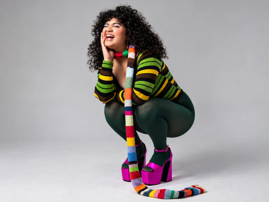 Remi Wolf crouches and laughs against a plain grey backdrop, wearing a green-striped knit top, dark green tights, hot pink platform heels and a long multicoloured striped scarf.