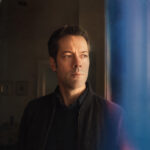 Robert Hinrichs gazes out of frame in a black bomber jacket, lit by warm window light against a blue-toned backdrop, in a promotional portrait for his single ‘The Road.’
