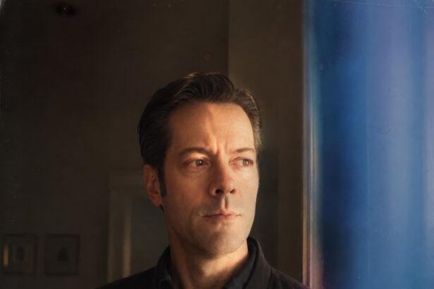Robert Hinrichs gazes out of frame in a black bomber jacket, lit by warm window light against a blue-toned backdrop, in a promotional portrait for his single ‘The Road.’
