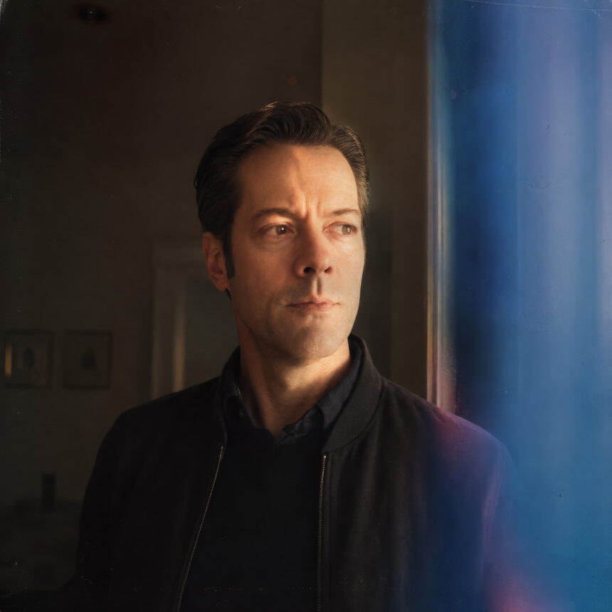 Robert Hinrichs gazes out of frame in a black bomber jacket, lit by warm window light against a blue-toned backdrop, in a promotional portrait for his single ‘The Road.’