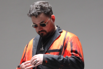 STRAiNGER looks down as he adjusts his sleeve, wearing hexagonal gold-rimmed sunglasses and an orange and black painted-print blazer against a plain grey backdrop.