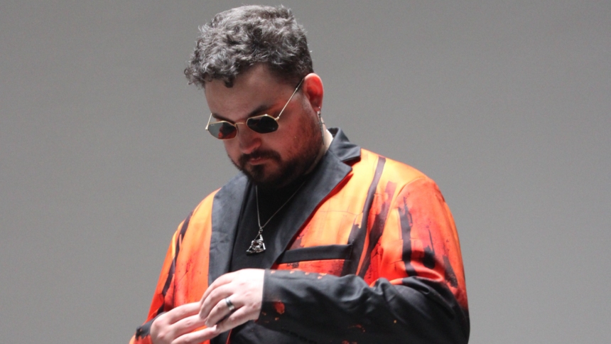 STRAiNGER looks down as he adjusts his sleeve, wearing hexagonal gold-rimmed sunglasses and an orange and black painted-print blazer against a plain grey backdrop.