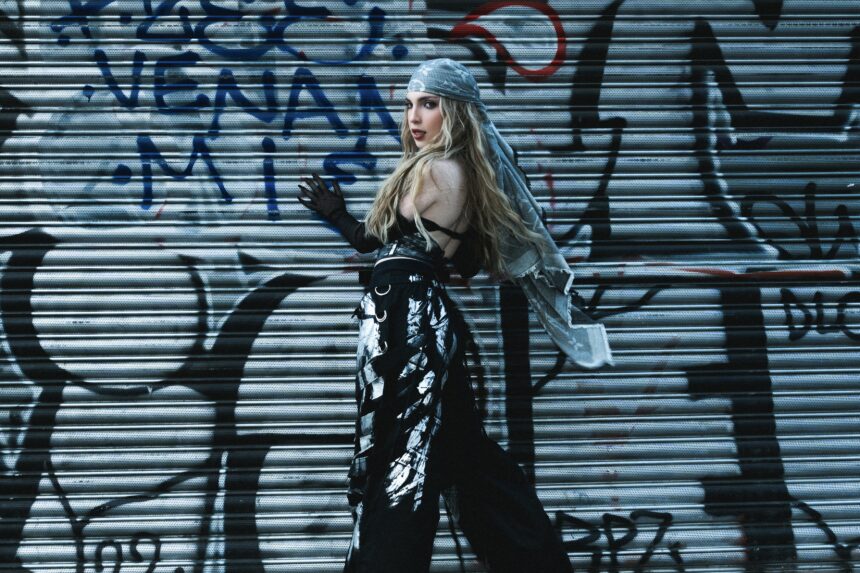 Pop artist Samsara looks over her shoulder against a graffiti-covered metal shutter, wearing a flowing grey headscarf, sheer black gloves and silver-painted black trousers.