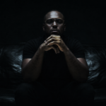 ScHoolboy Q sits on a dark couch in near-black, moody lighting, hands clasped in front of his mouth, wearing a dark short-sleeve shirt and silver rings.