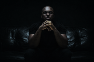 ScHoolboy Q sits on a dark couch in near-black, moody lighting, hands clasped in front of his mouth, wearing a dark short-sleeve shirt and silver rings.