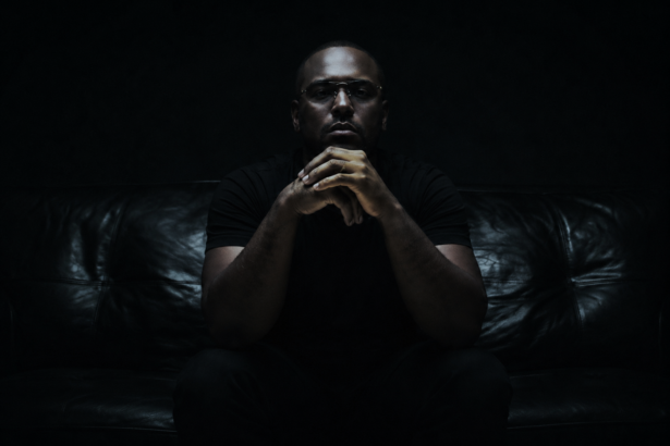ScHoolboy Q sits on a dark couch in near-black, moody lighting, hands clasped in front of his mouth, wearing a dark short-sleeve shirt and silver rings.