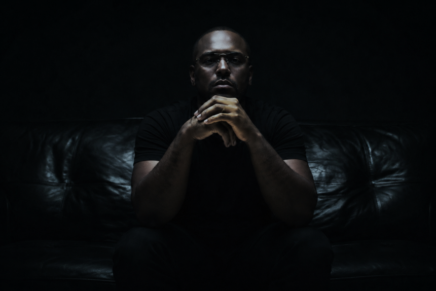 ScHoolboy Q sits on a dark couch in near-black, moody lighting, hands clasped in front of his mouth, wearing a dark short-sleeve shirt and silver rings.