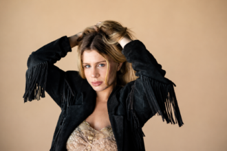 Slayyyter runs her hands through tousled blonde hair, wearing a black suede fringe jacket over a gold embellished lace top against a warm peach backdrop.