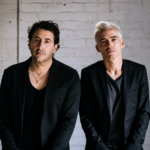 Two members of The Avalanches stand side by side against a whitewashed brick wall, both in black blazers and dark V-neck tops.