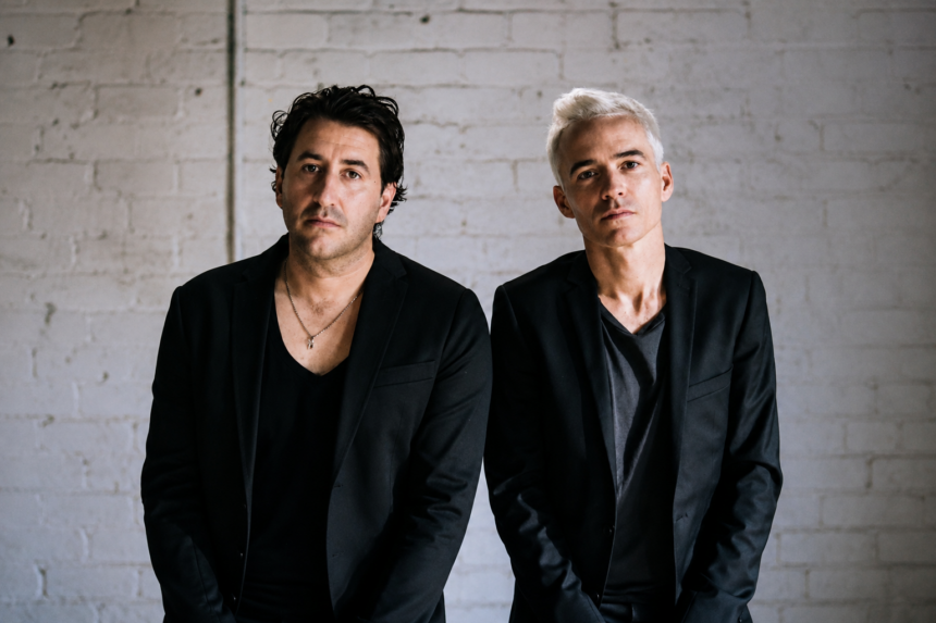 Two members of The Avalanches stand side by side against a whitewashed brick wall, both in black blazers and dark V-neck tops.