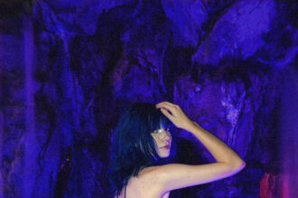 Alt-pop artist Wormogenic looks over her shoulder in a glittering dark tulle dress, standing in glowing blue water before a purple-lit rock grotto.