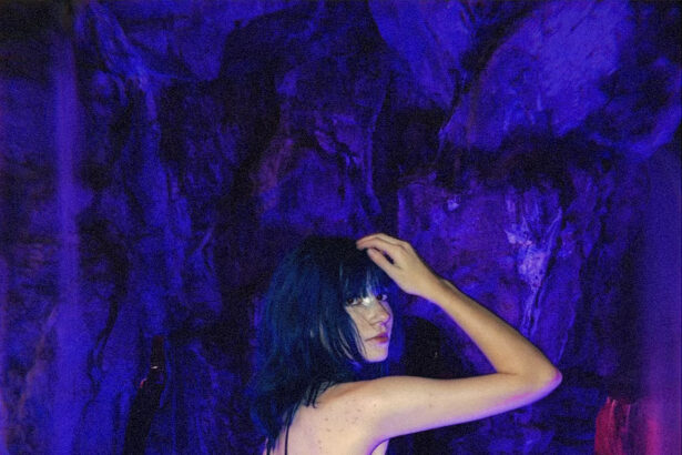 Alt-pop artist Wormogenic looks over her shoulder in a glittering dark tulle dress, standing in glowing blue water before a purple-lit rock grotto.