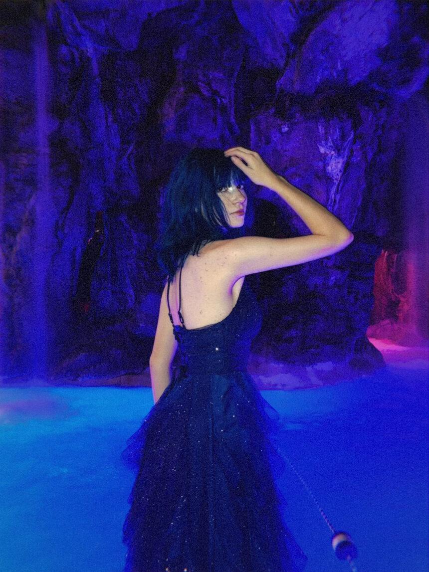 Alt-pop artist Wormogenic looks over her shoulder in a glittering dark tulle dress, standing in glowing blue water before a purple-lit rock grotto.
